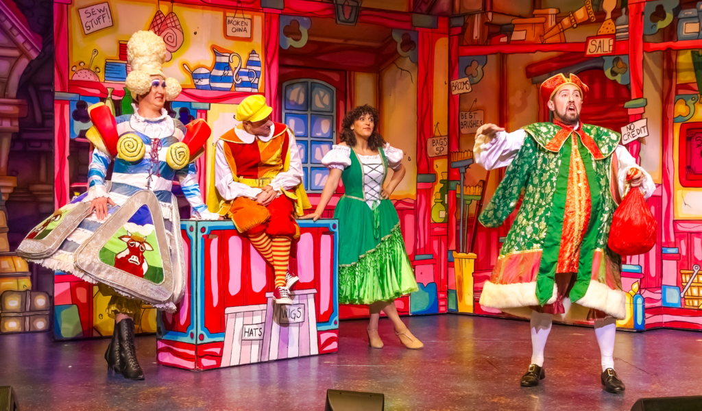 Dick Whittington Production Photo: 4 characters in a colourful shop; Sarah Suet (Matt Freeman) is leaning against the counter in a dress decorated with cheese triangles; Jack (Al Dunn) is sitting on the counter in an orange and yellow outfit; Alice (Sophia Lewis) is leaning on the other side of the counter in a green dress, with one hand on her hip; they are all watching Alderman Fitzwarren (Nick Bunt) who stands in the foreground in fine gold-trimmed clothes, gesturing with one hand and clutching a red sack in the other.