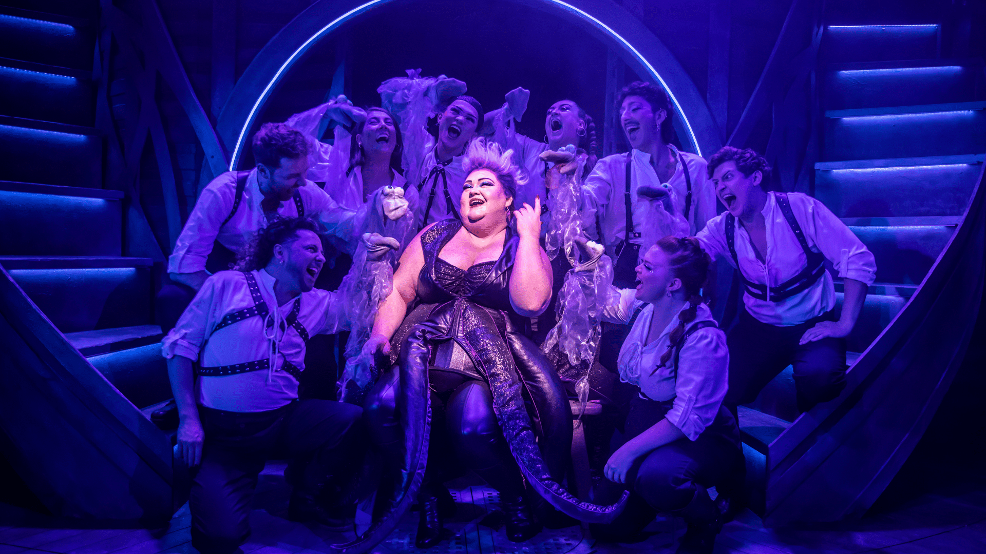 Unfortunate Production Photo. Ursula sits in a pink spotlight wearing a scaly, black bustier with tentacles attached. She is surrounded by other performers in white shirts and black leather harness. All are singing.