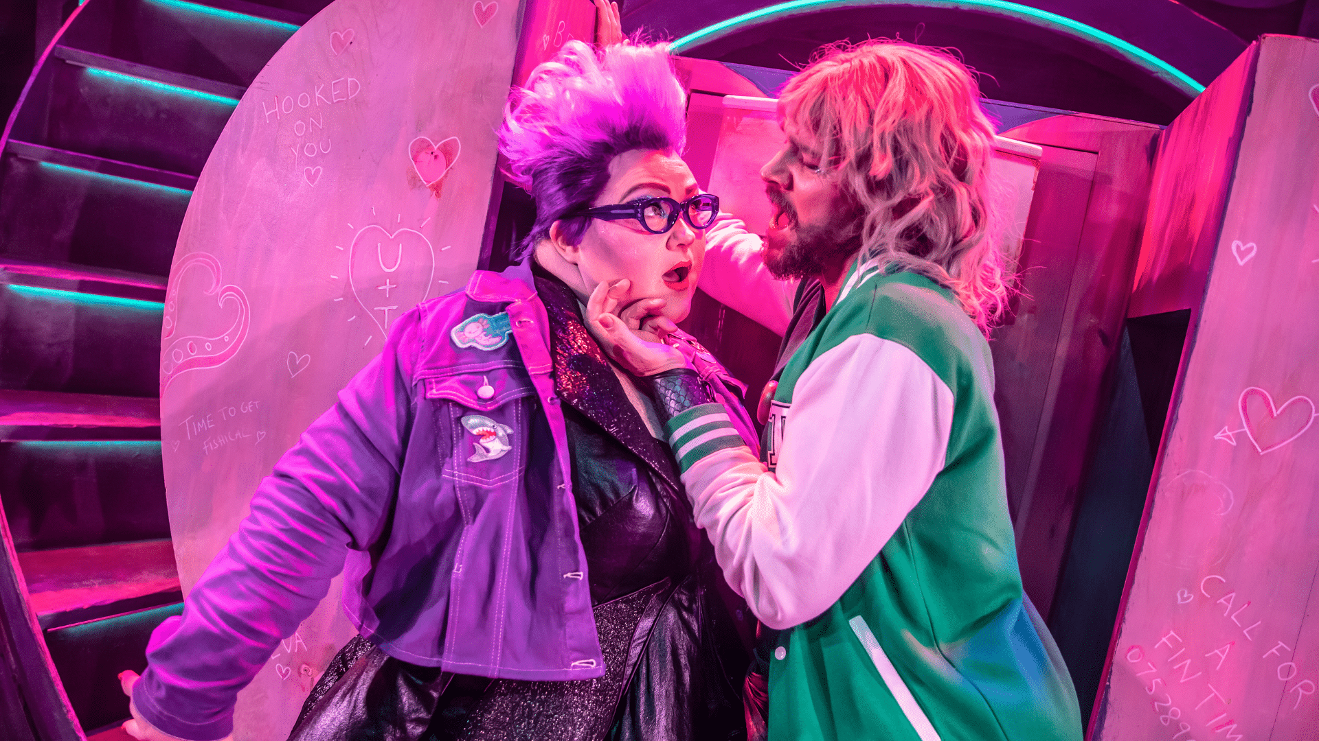 Unfortunate production photo. Ursula (wearing glasses and a denim jacket) looks into the eyes of a man with shoulder-length blonde hair and a green varsity jacket, who is stroking her cheek. Both are singing.