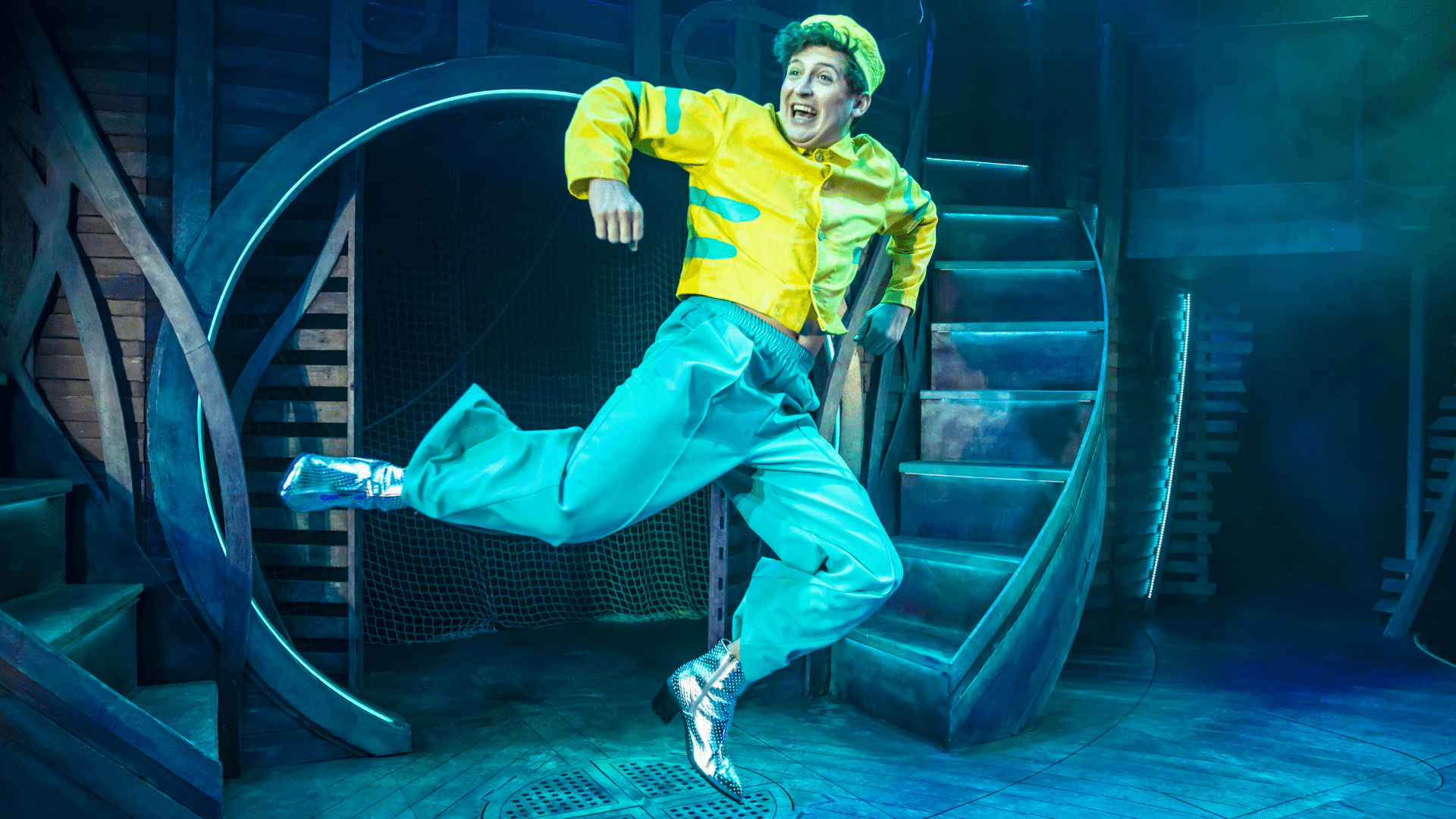 Unfortunate production photo. A blue, nautical stage-set with stairs curving upwards either side of a circular doorway. A performer in a yellow hi-vis jacket and hat leaps into the air.