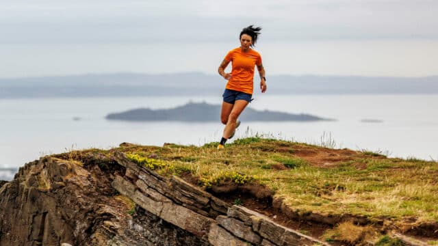 Allie Bailey, a middle-aged white woman with long, tied back black hair, wearing an orange running top and blue shorts, runs on the edge of a grassy cliff along a coastline. Behind her, an island in the ocean, and hills in the distance beyond that.