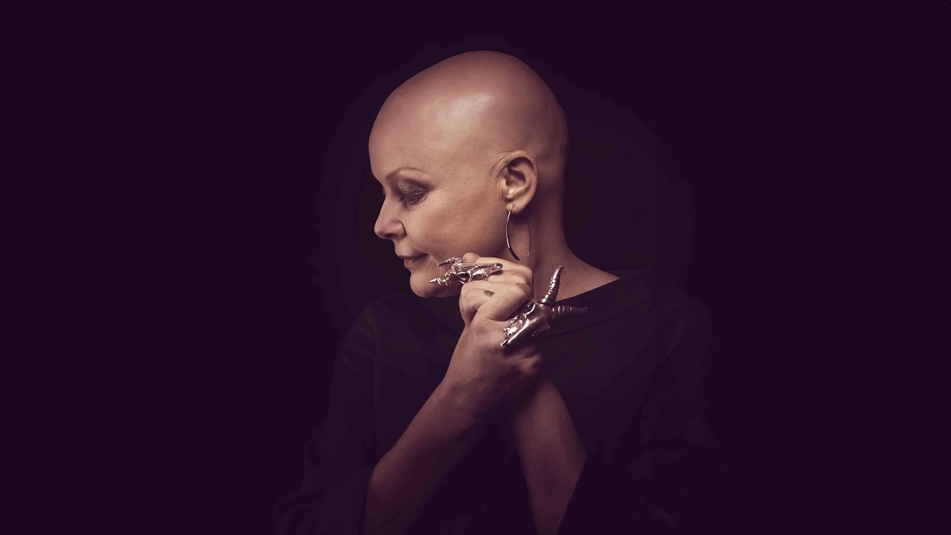 Black background. Gail Porter, a middle aged white woman with alopecia, faces the camera with her body but looks over her right shoulder, clasping her hands together. She is wearing a dark purple top, and sheep skull-shaped rings on her middle fingers