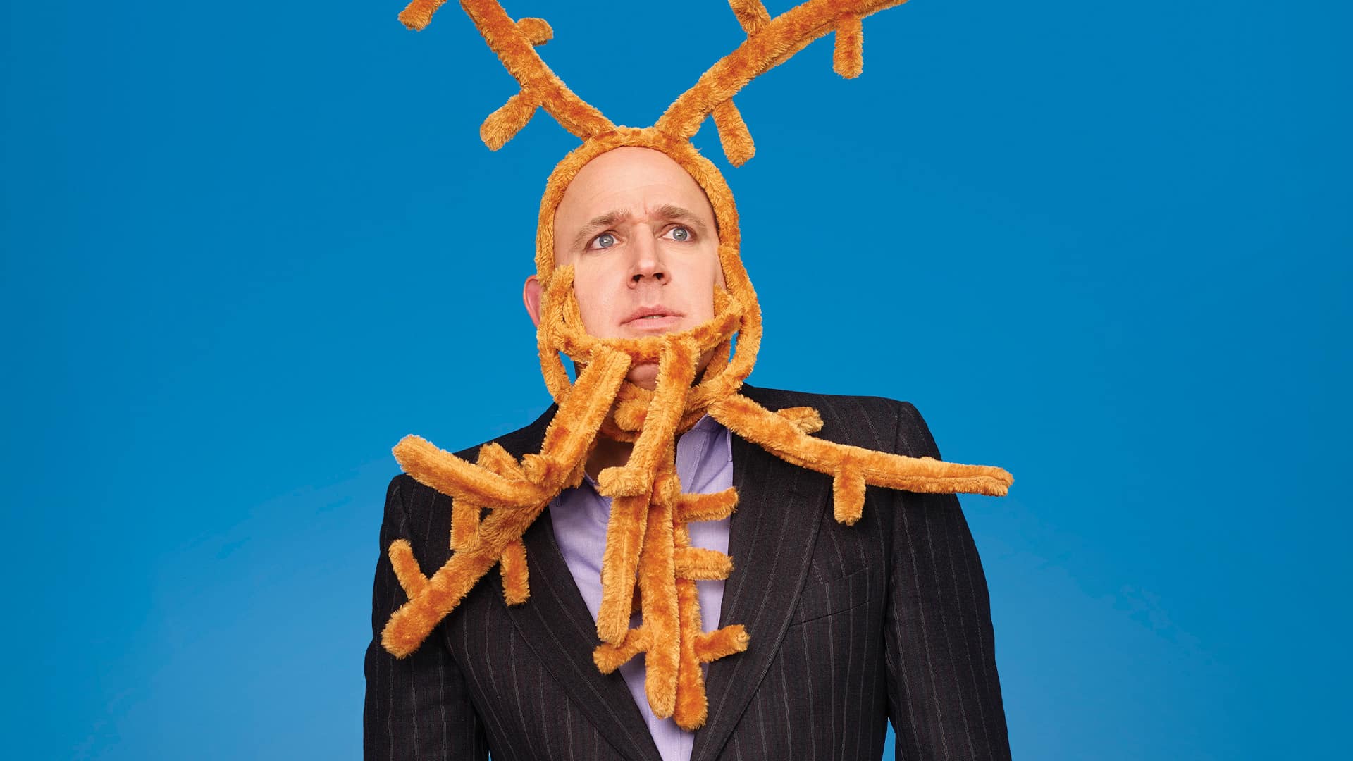 Tim Vine: Breeeep! artwork. Blue background. Tim Vine, wearing multiple toy reindeer antlers around his face, and a pinstripe suit over a light grey / pink shirt, looks off camera with a confused expression.
