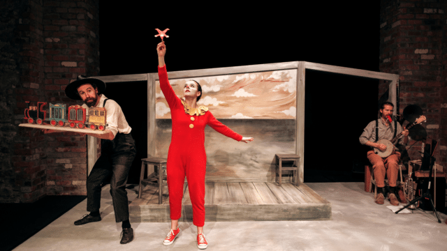 The Farmer & the Clown production photo. A bare stage aside from a dusty wooden platform and a painted backdrop showing a sepia-toned flat landscape. A performer dressed in a red onesie with yellow buttons and a ruffled comma holds a small cut-out clown, similarly dressed, upside-down in the air. Another performer, dressed in a wide-brimmed black hat holds a hand-painted model train.