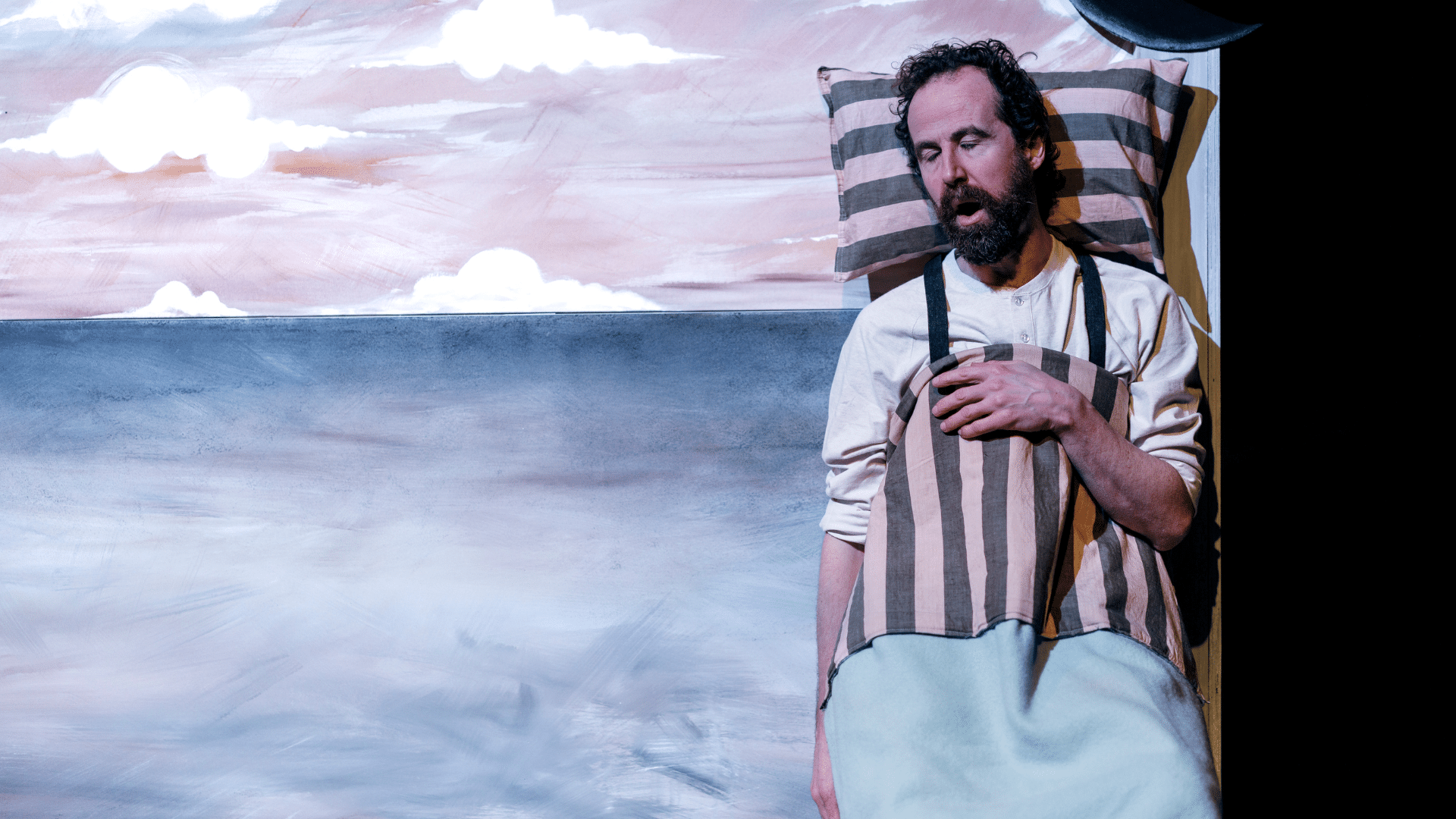The Farmer & the Clown production photo. A bearded performer stands against a painted backdrop of a barren landscape. His head is resting on a striped pillow, and he holds a matching eiderdown to his chest, as if sleeping.