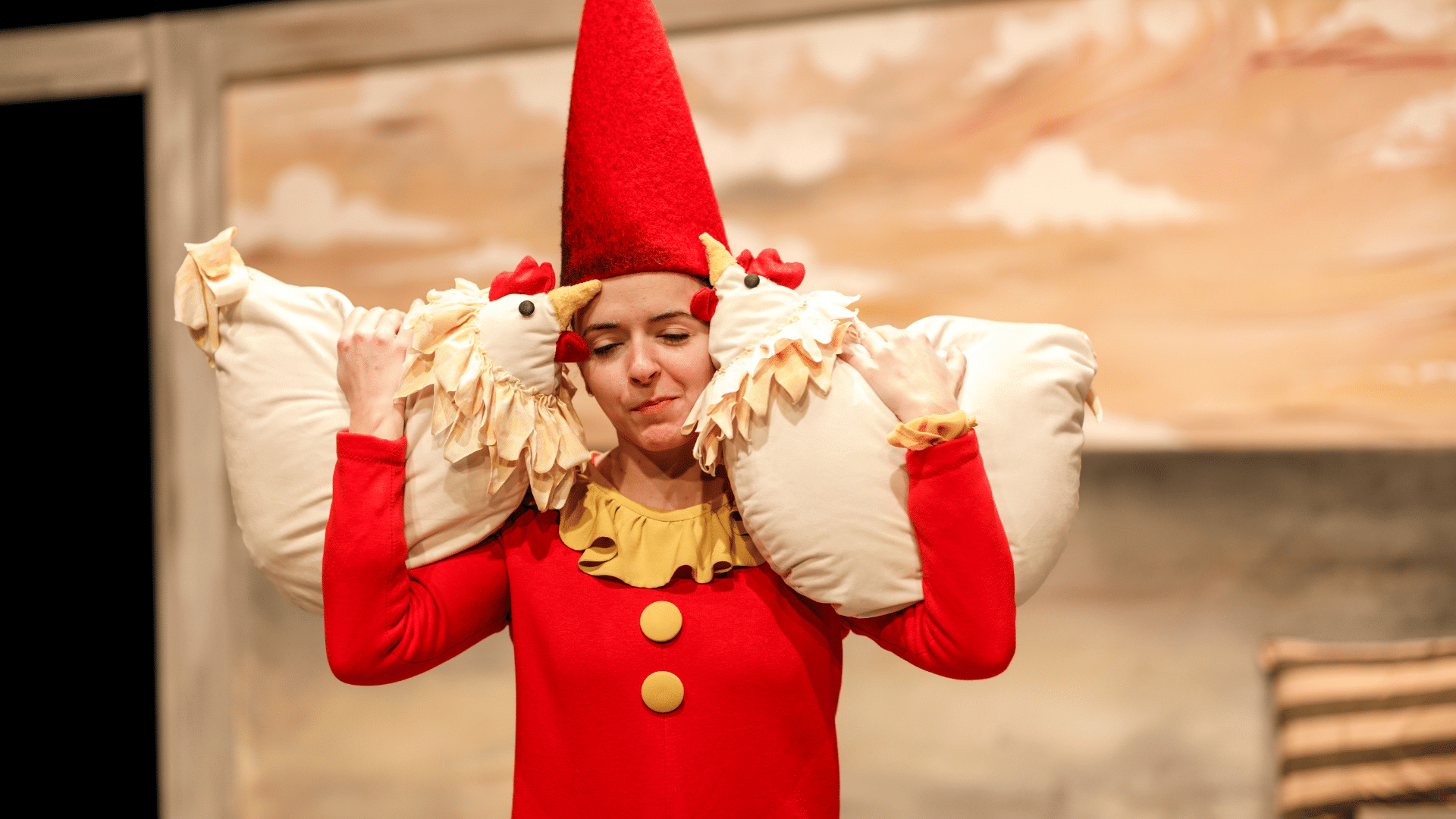 The Farmer & the Clown production photo. A performer in a conical scarlet hat and matching onesie holds two stuffed chickens on their shoulders. They close their eyes and smile slightly.