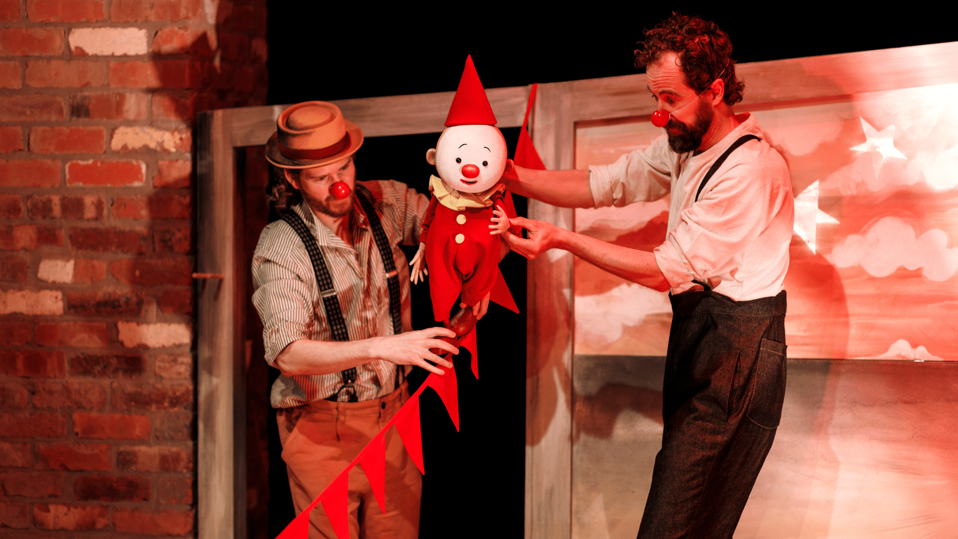 The Farmer & the Clown production photo. Two performers wearing shirts and red noses puppeteer a small clown dressed in red. The puppet appears to be tightrope walking along scarlet bunting.