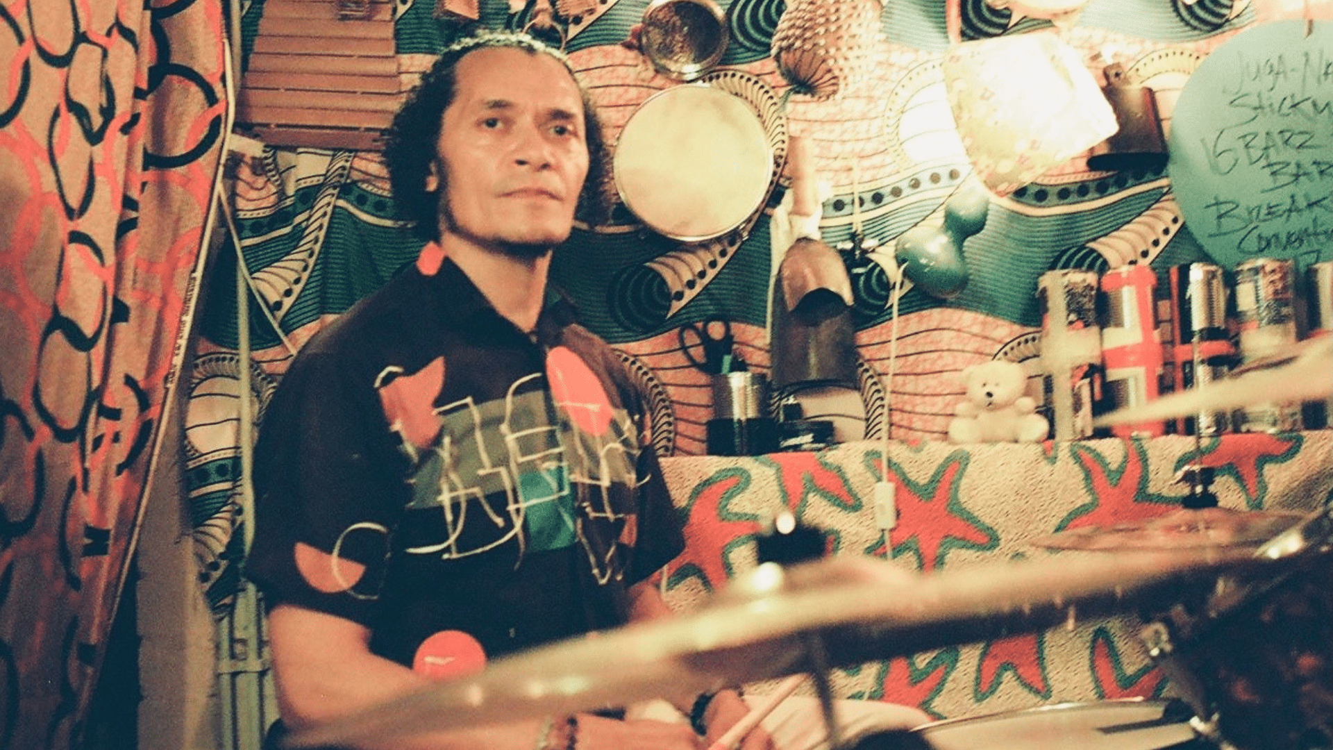 Photo of David Stickman Higgins in his studio. He's a Carribean man with mid-long curly hair. On the wall are many colourful and patterned fabrics and a variety of percussion instruments.
