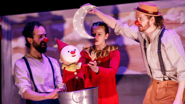 A woman dressed in a red onesie controls a sweet looking baby clown puppet, which looks up at a light up crescent moon being held by another actor.