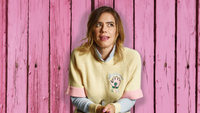 Photo. Pink wooden-panelled background. Lou Sanders, wearing a pastel-coloured knitted jumper, half-smiles and casts her gaze to the side.