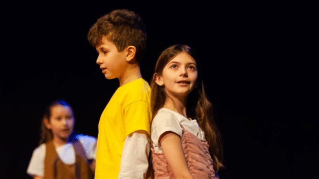 Two children (a boy wearing a yellow t-short and a girl in a pink dress) stand back to back under a bright spotlight. The background is black theatre curtains.