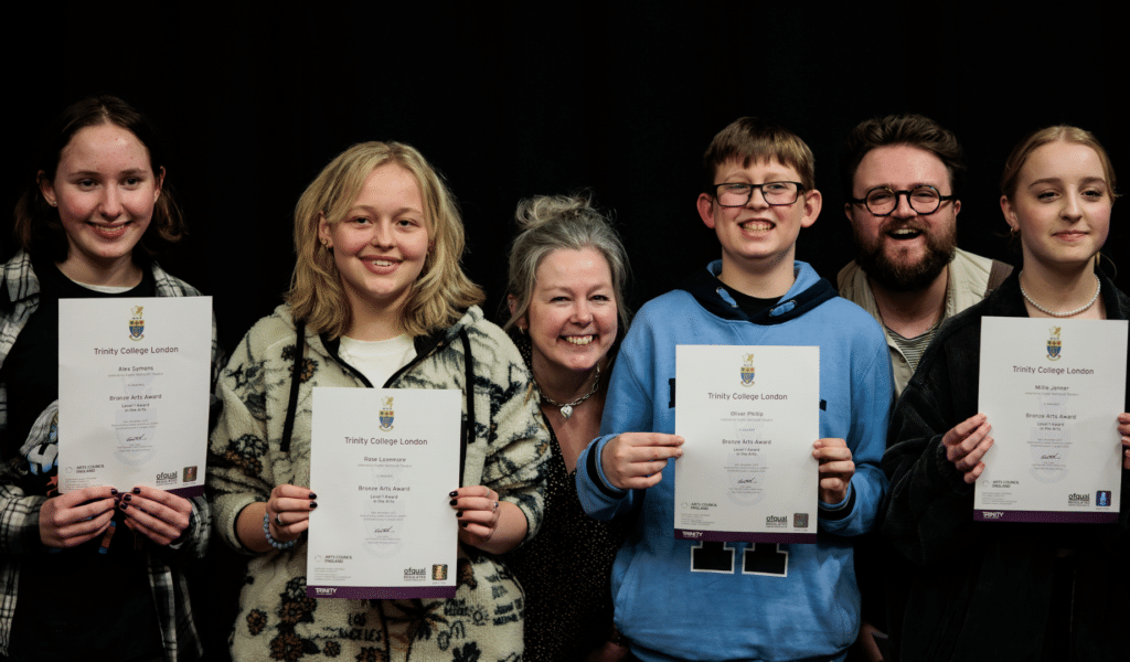 A photo of four young people standing side-by-side, smiling. Each holds a certificate in front of them. Two adults photobomb from behind.