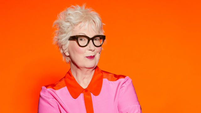 Jenny Eclair is standing against a bright orange background. He's giving the camera a cheeky glance and smile. She's wearing a pink blouse with orange collar and thick black-rimmed glasses.