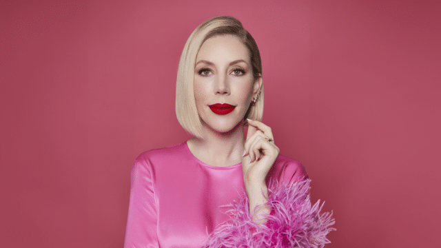 Katherine Ryan is dressed in a bright pink silk top with a feathery sleeve. Her blonde hair is cut in a long bob and she's wearing her signature red lipstick.