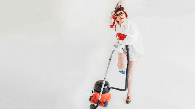 Laura Belbin is wearing an oversized white shirt and one shoe. She's trying to hold her other shoe, a pair of sunglasses, on old-fashioned red phone, a kettle and the piping of a red Henry hoover to comical effect.