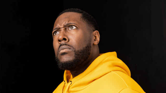 The comedian Aurie Styla wearing a bright yellow hoodie with a black background behind him.