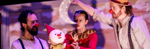 A woman dressed in a red onesie controls a sweet looking baby clown puppet, which looks up at a light up crescent moon being held by another actor.