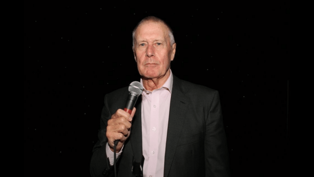 Black background. Geoff Hurst, a balding man in a black blazer and pale pink shirt, holds a microphone to his stoic mouth.