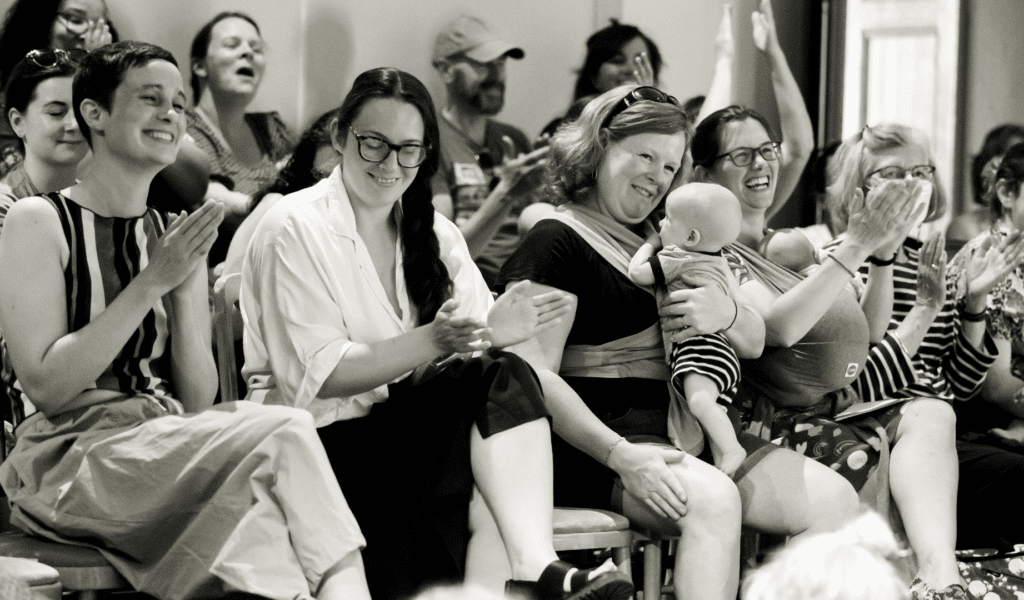 A black and white photo of people smiling and applauding while sitting in auditorium seats. Some of the people are holding small children/babies on their laps, or carrying babies in slings.