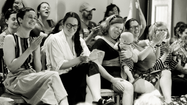 A black and white photo of people smiling and applauding while sitting in auditorium seats. Some of the people are holding small children/babies on their laps, or carrying babies in slings.