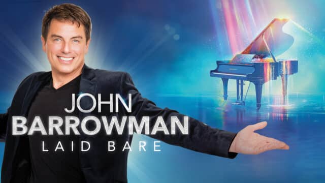 John Barrowman Laid Bare: John is wearing a black suit and is looking into the camera with open arms and a broad smile. Behind him is a piano covered in blue and pink light and lots of sparkle.