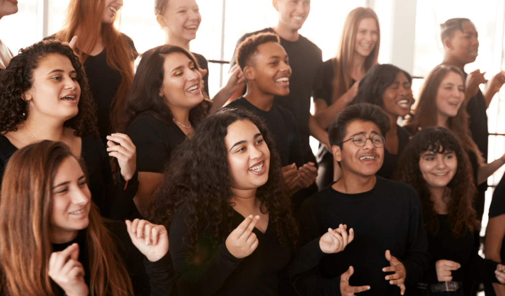 A photo of a diverse group of young people singing together. They are all wearing black and smiling. Sunlight streams through a window behind them.