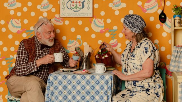 An elderly couple wearing smart but eccentric clothing sit at a small table covered in a gingham table cloth for afternoon tea. To the right is the edge of a kitchen dresser. The wall behind them is covered in orange wallpaper, patterned with tea pots.