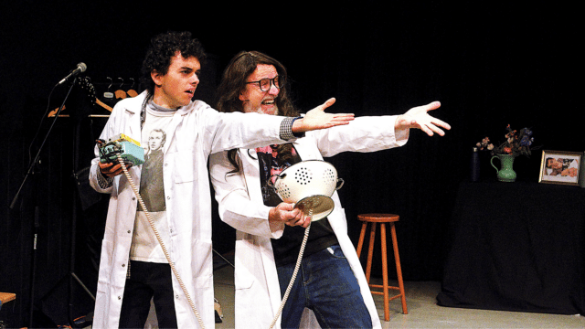 Washing Machine of Destiny Production Photo. Two performers wearing white lab-coats grip distinctly homemade equipment (I.e. a colander wired into the machinery) and gesture at something off-camera.