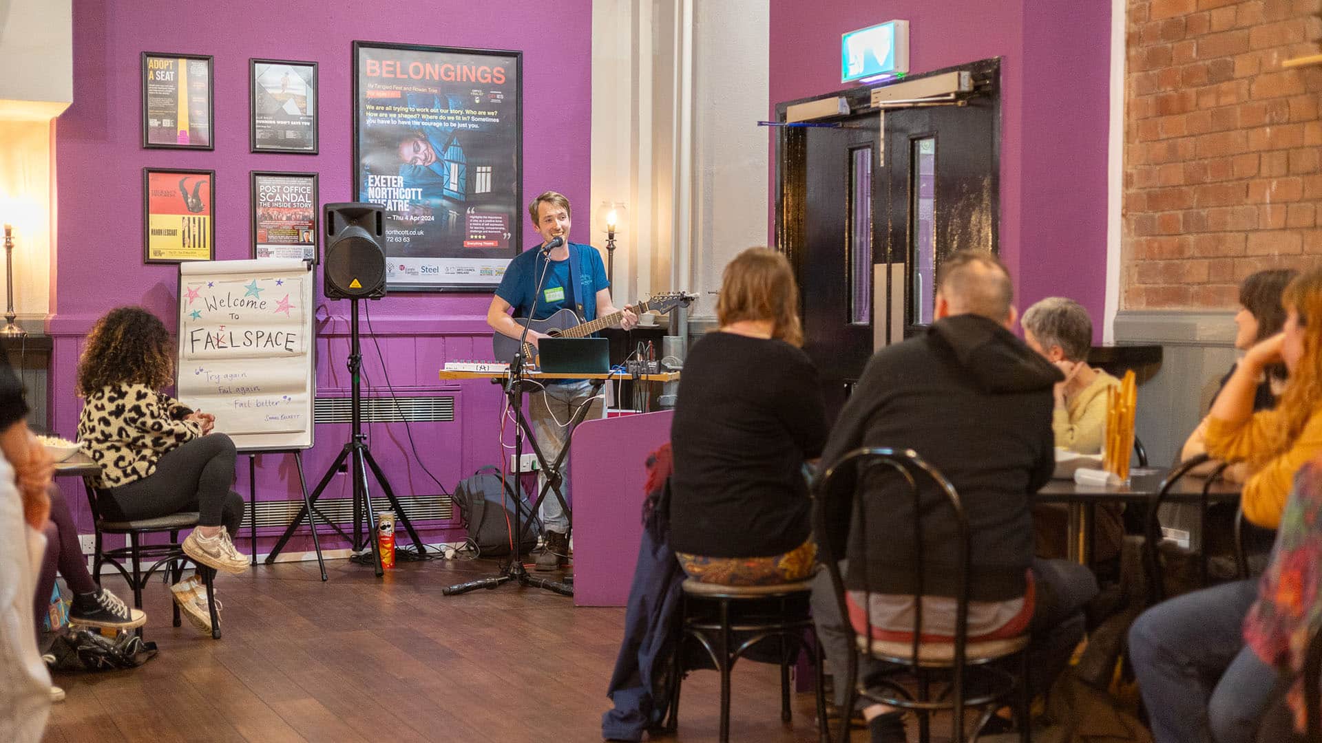 A musician with an acoustic guitar is playing music for a group of Failspace participants sat at tables in the Exeter Barnfield Theatre's bar.