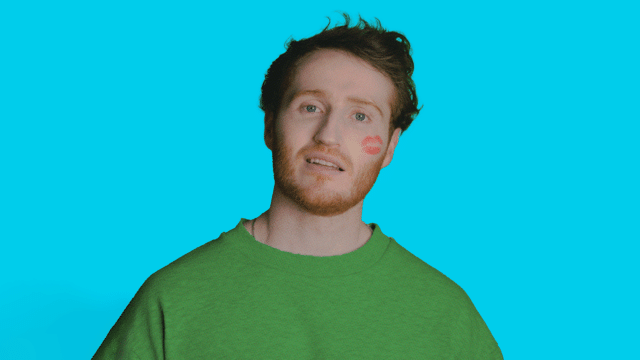A photo of Ali Woods with a love -struck, slightly befuddled look on his face, the imprint of a lipstick-red kiss on his cheek. He's wearing a bright green sweater and the background is a bright blue.