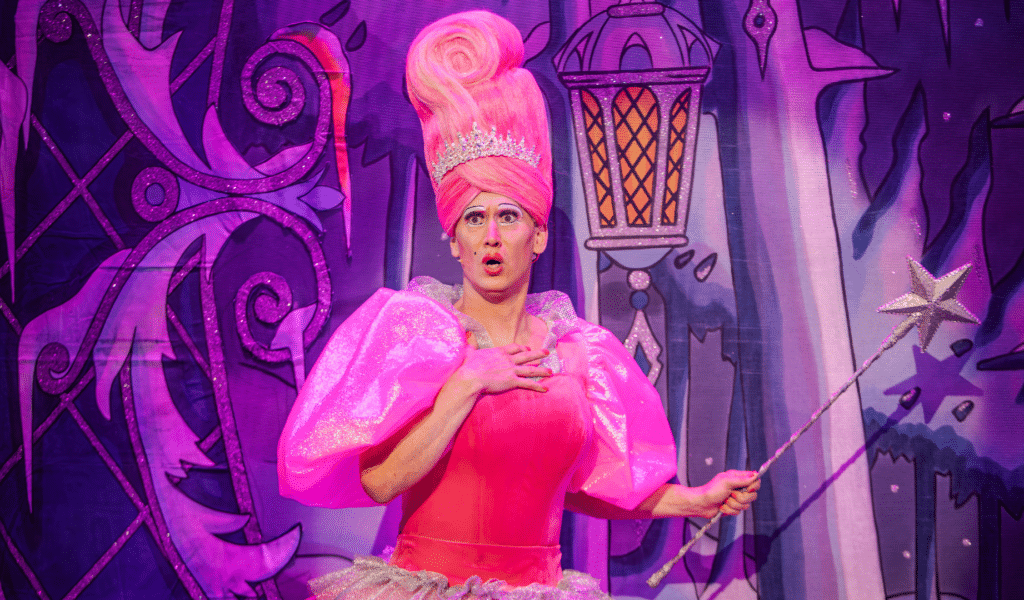 Photo of the Fairy Godmother, wearing a pink dress with enormously puffy sleeves and a matching, towering wig. She puckers, holds a hand to her breast, and clutches a silver, star-topped wand with the other.