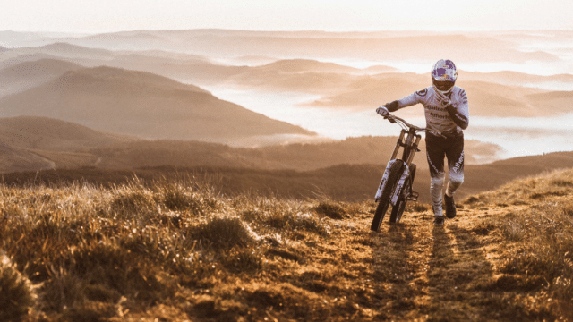 Gee Atherton wearing biking gear and a helmet, pushing his bike through a mountainous landscape.