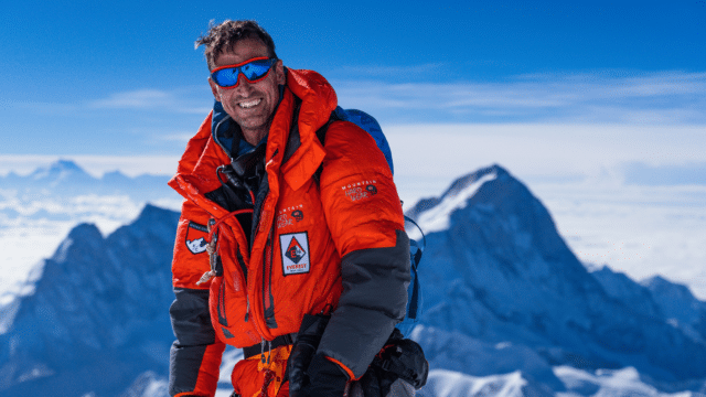 Kenton Cool stands in front of a snowy mountainous landscape, smiling, wearing orange climbing gear and goggles.