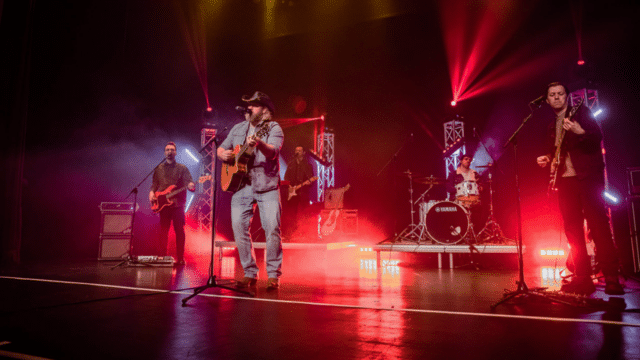 Male modern country music singer performs with a band under red stage lighting.
