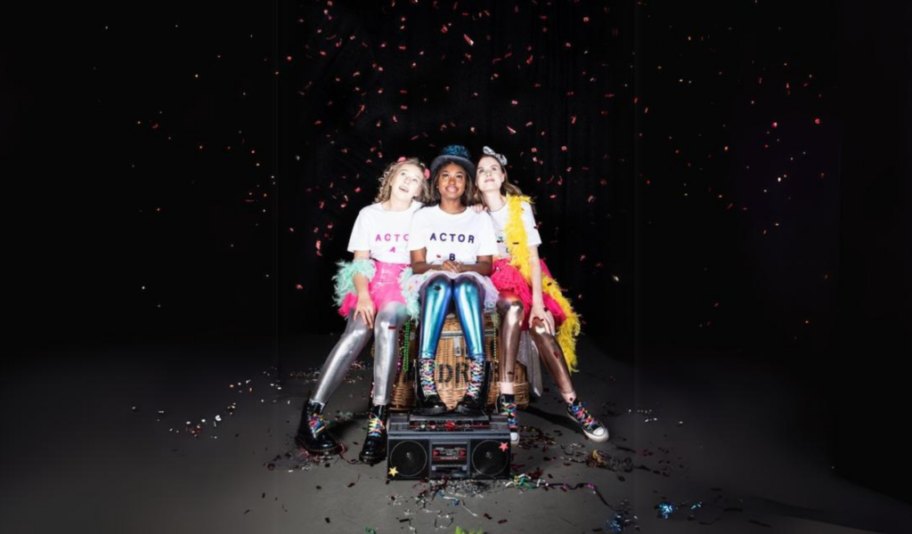 Artwork for Bright Places: Three women dressed in feather boas, bright leggings, and T-shirts that say 'Actor' are sat in the spotlight on a dark stage with a jukebox at their feet. Confetti falls from above and they look up, smiling.