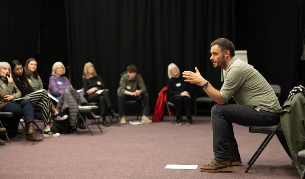 A photo of Martin Berry, wearing a shirt and jeans, leading a conversation of people seated on chairs in a rehearsal studio, facing him.
