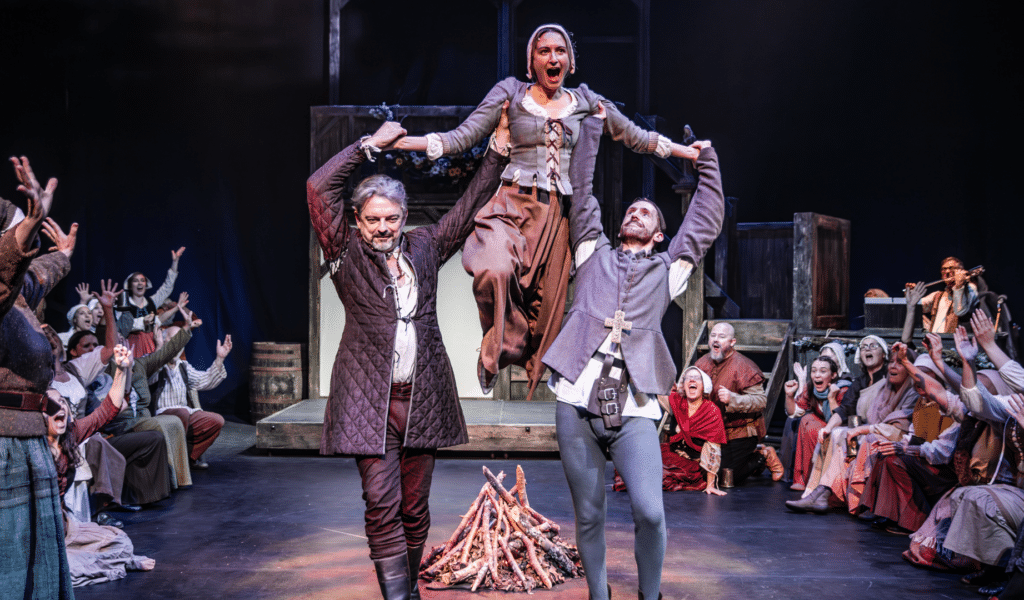 Photo of a woman being lifted over a fire by two men. They are surrounded by cheering ensemble members. All are in medieval dress and look joyful.