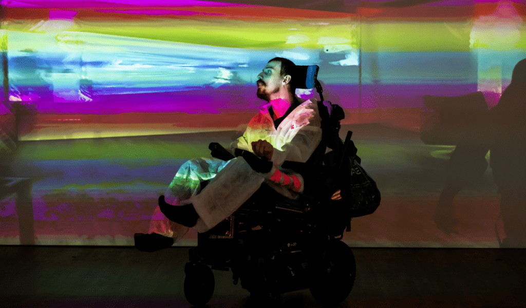 Centre: Hugh Malyon, a performer with close-cropped dark hair and a moustache and beard in a wheelchair, in front of a wall displaying a blur of rainbow colours.