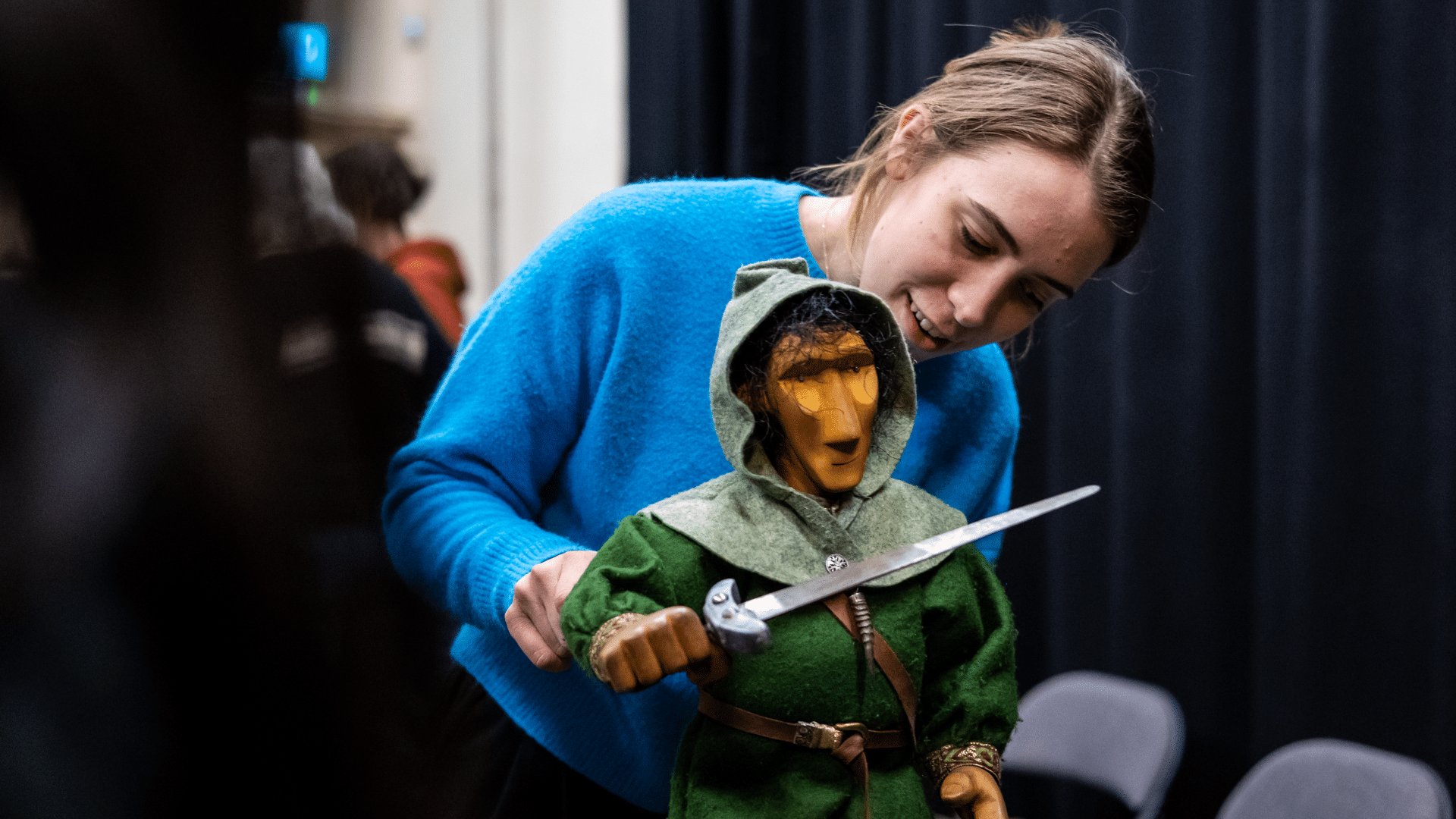 A young woman in a blue jumper operates an intricate puppet knight, her face serene with concentration.