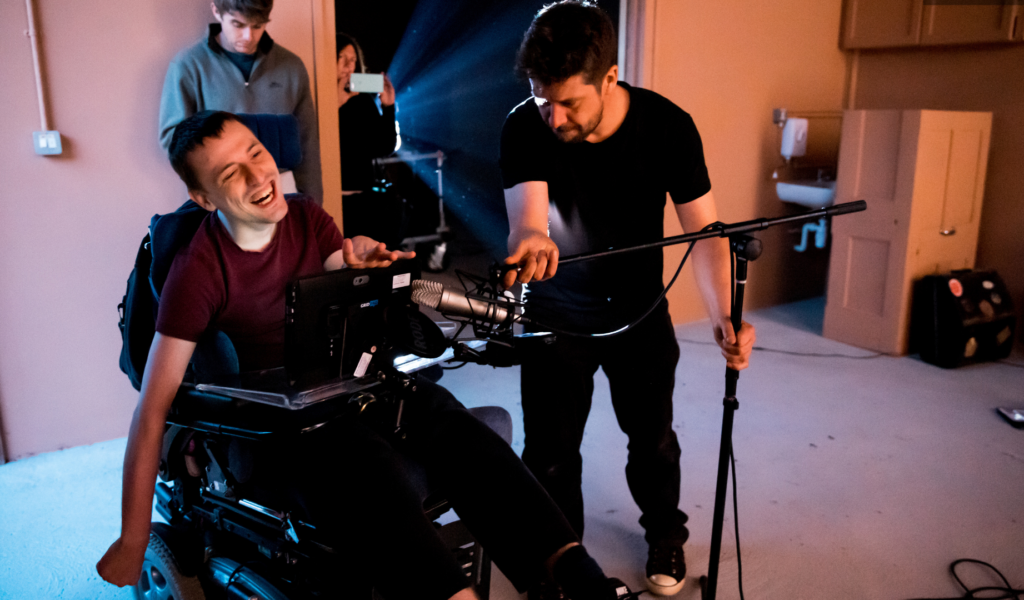 Hugh Maylon smiles while another theatre maker sets up a microphone in front of him.