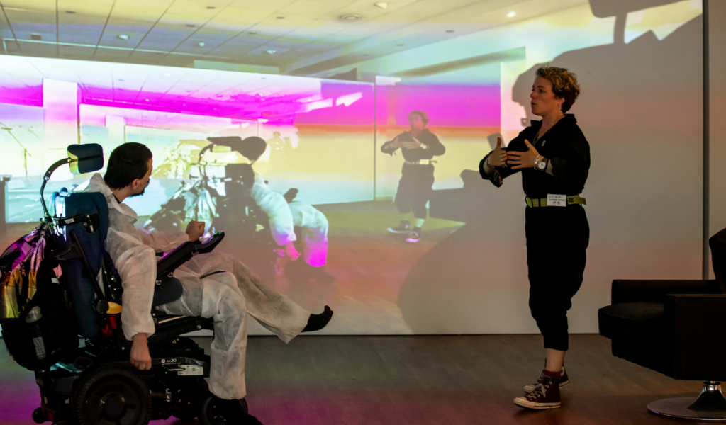 Two performers in a studio space in front of a white wall with a projected spectrum of neon colours on it.