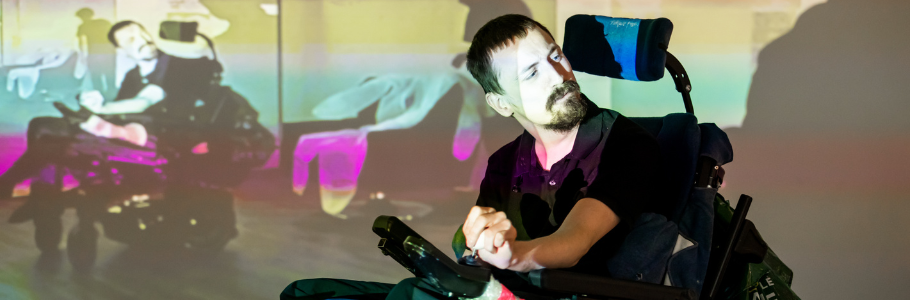 Centre: Hugh Malyon, a performer with close-cropped dark hair and a moustache and beard in a wheelchair, in front of a wall displaying a blur of rainbow colours with himself projected among them.