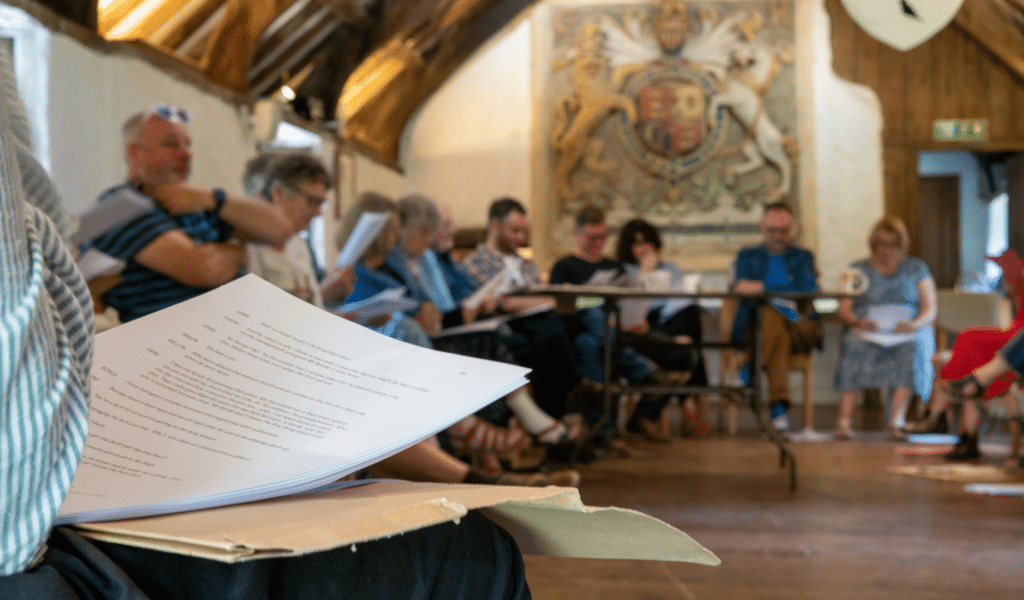 A script is seen in the foreground, on the lap of a man in a pinstriped shirt. In the background, a circle of seated people in a vaulted hall, each with a script on their lap.