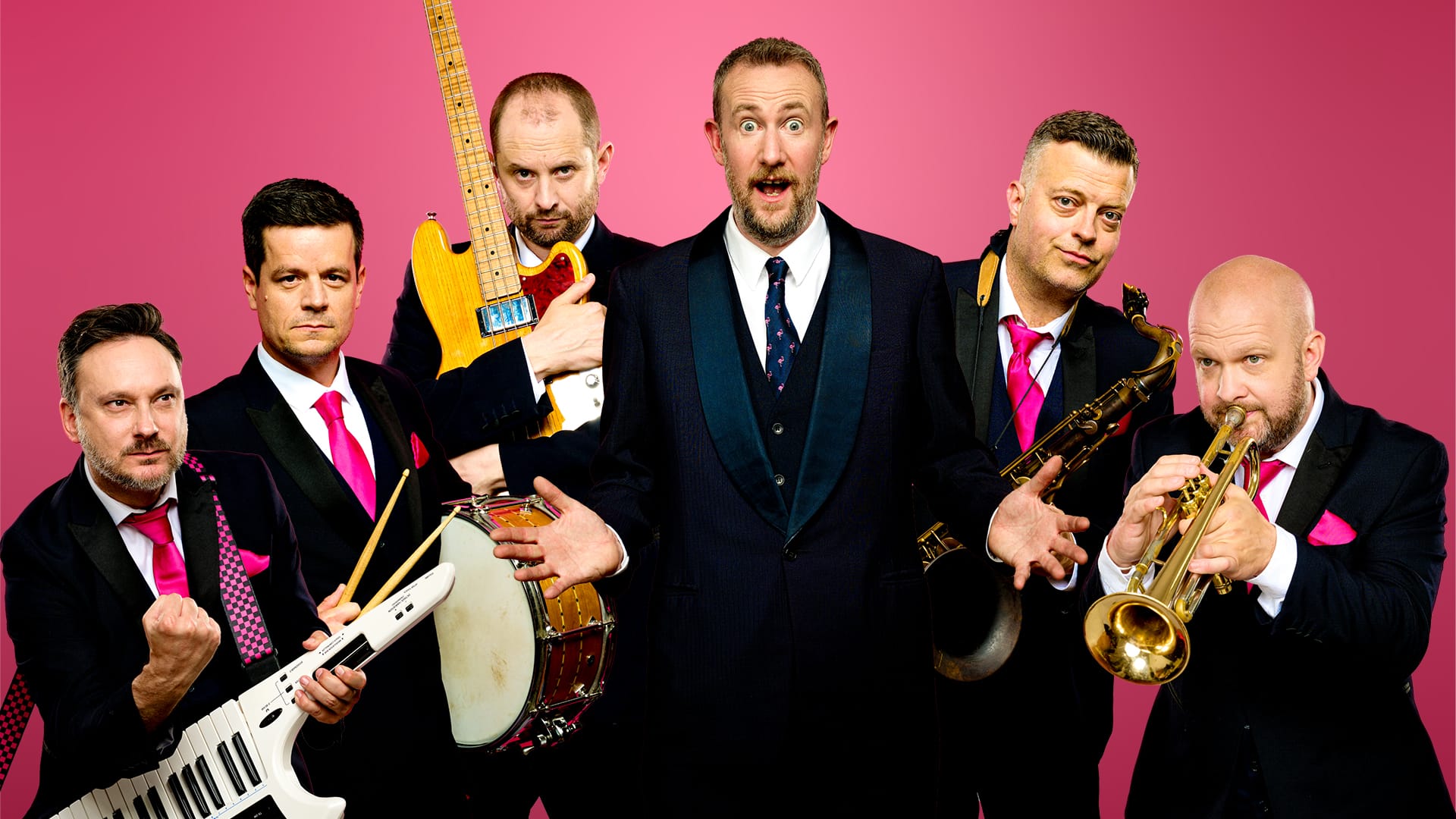 The full Horne Section band, with Alex Horne in the centre are holding their instruments and making excited and comedic faces to the camera. They're wearing black blazers and pink ties.
