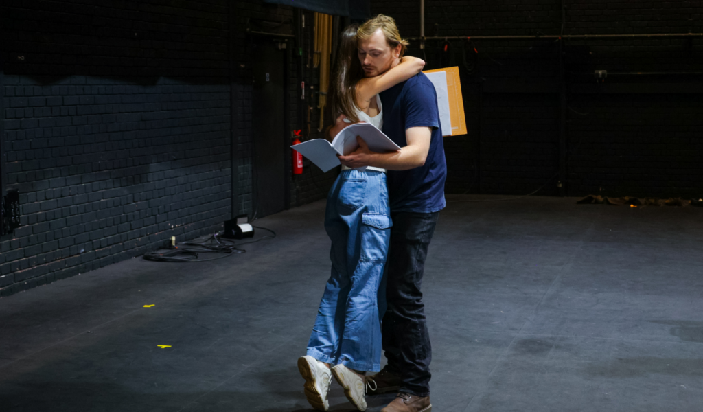 Two casually-dressed actors holding scripts hug in a black rehearsal space.