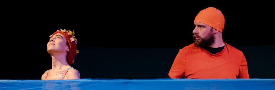 Photograph of two actors stood behind a shoulder-height blue gauze, simulating water. On the left, a woman in a beanie decorated with flowers looks up to the sky serenely. On the right, a man in an orange rash vest and swimming cap looks at her intensely.