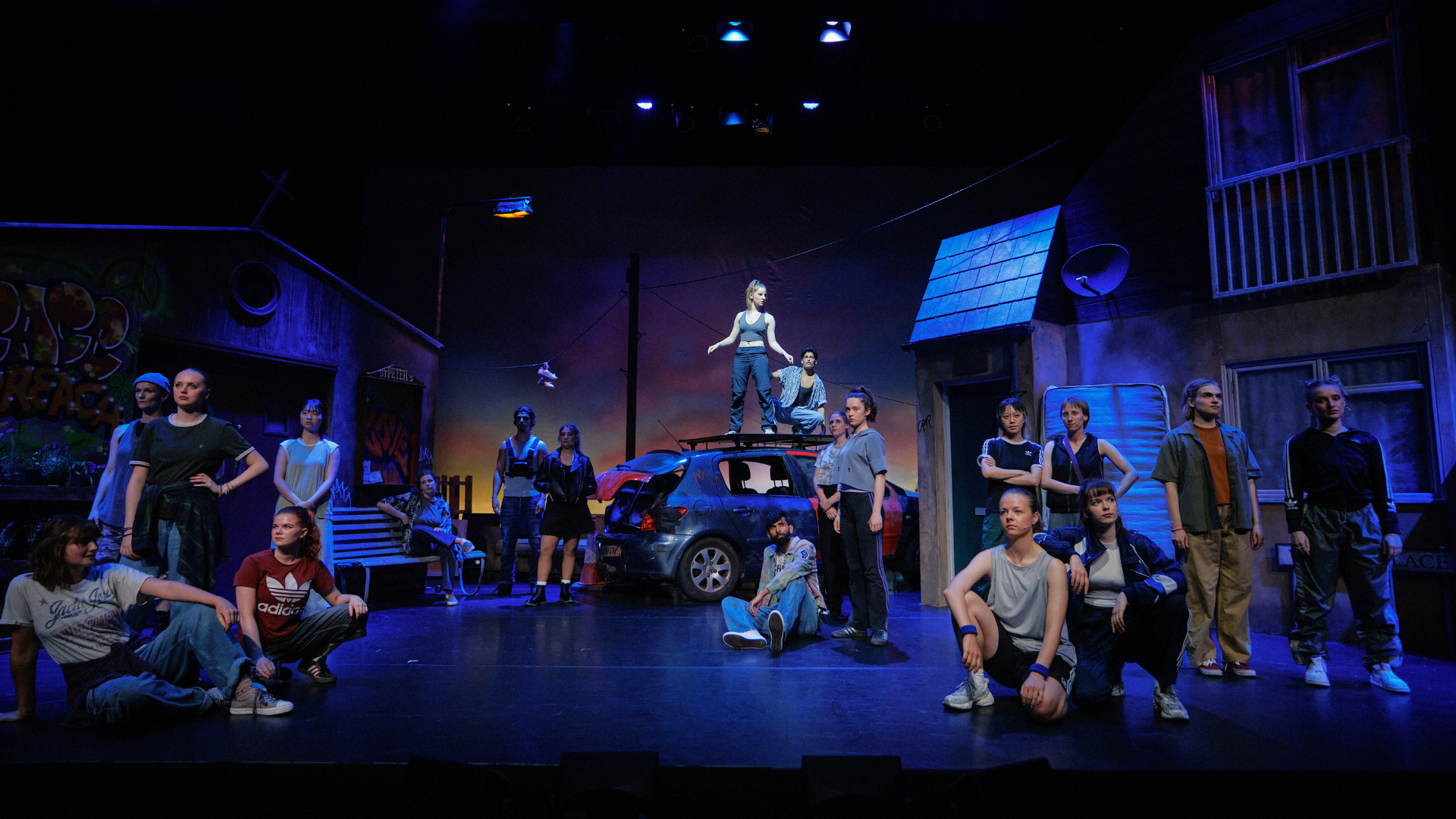 Photo of nineteen casually dressed young people scattered across a theatre set of an urban street. Two of them are stood on the roof of a battered car.