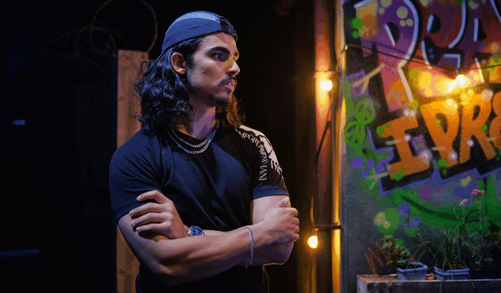 Photo of an actor in a black t-shirt with a black baseball cap (worn backwards) and a silver chain standing with arms crossed in front of colourful graffiti and a string of warm lightbulbs.