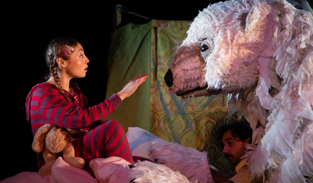 Photo of an awe-struck performer in stripy pink pyjamas face-to-face with a life-sized polar bear puppet, reaching her hand towards his nose.