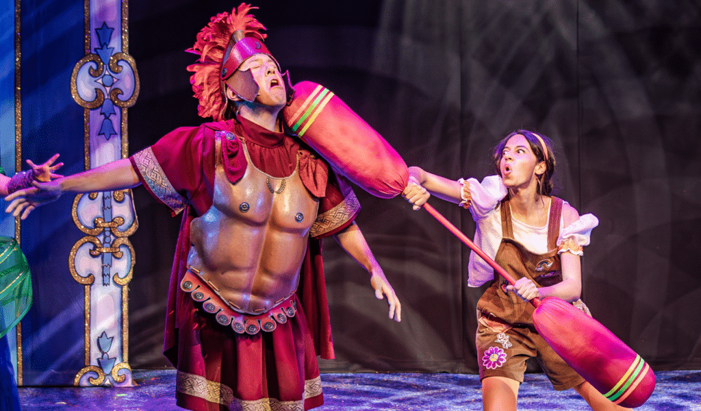 Photo of Cinderella, wearing brown overalls and orange tights, wields a pugel stick against a Roman gladiator.